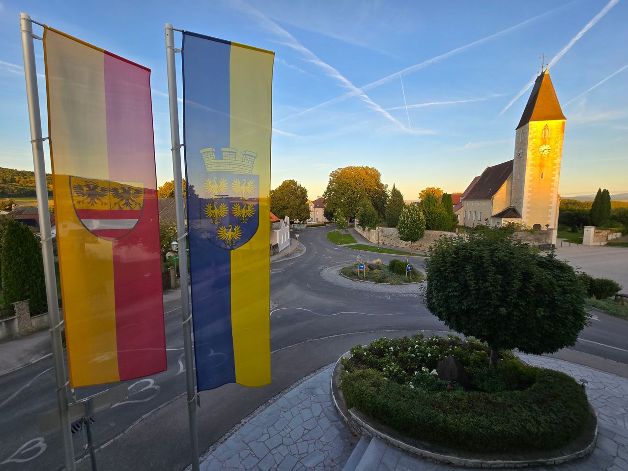 Eine Fahne mit einem Wappen und zwei weitere Fahnen befinden sich in der Nähe einer Straße. Eine Kirche mit Turm ist in der Ferne zu sehen. Bäume und Gebäude sind in der Umgebung. Der Himmel ist blau mit Wolken.