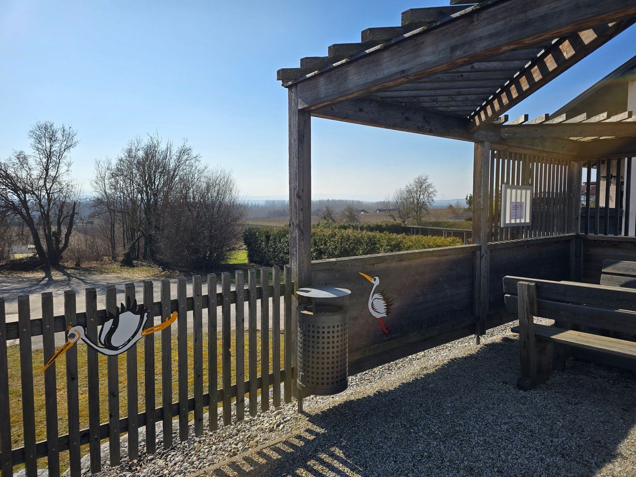 Ein hölzerner Pavillon mit einem Mülleimer und einer Bank vor einem Holzzaun mit einem Storch-Design. Im Hintergrund sind Bäume und Büsche zu sehen.