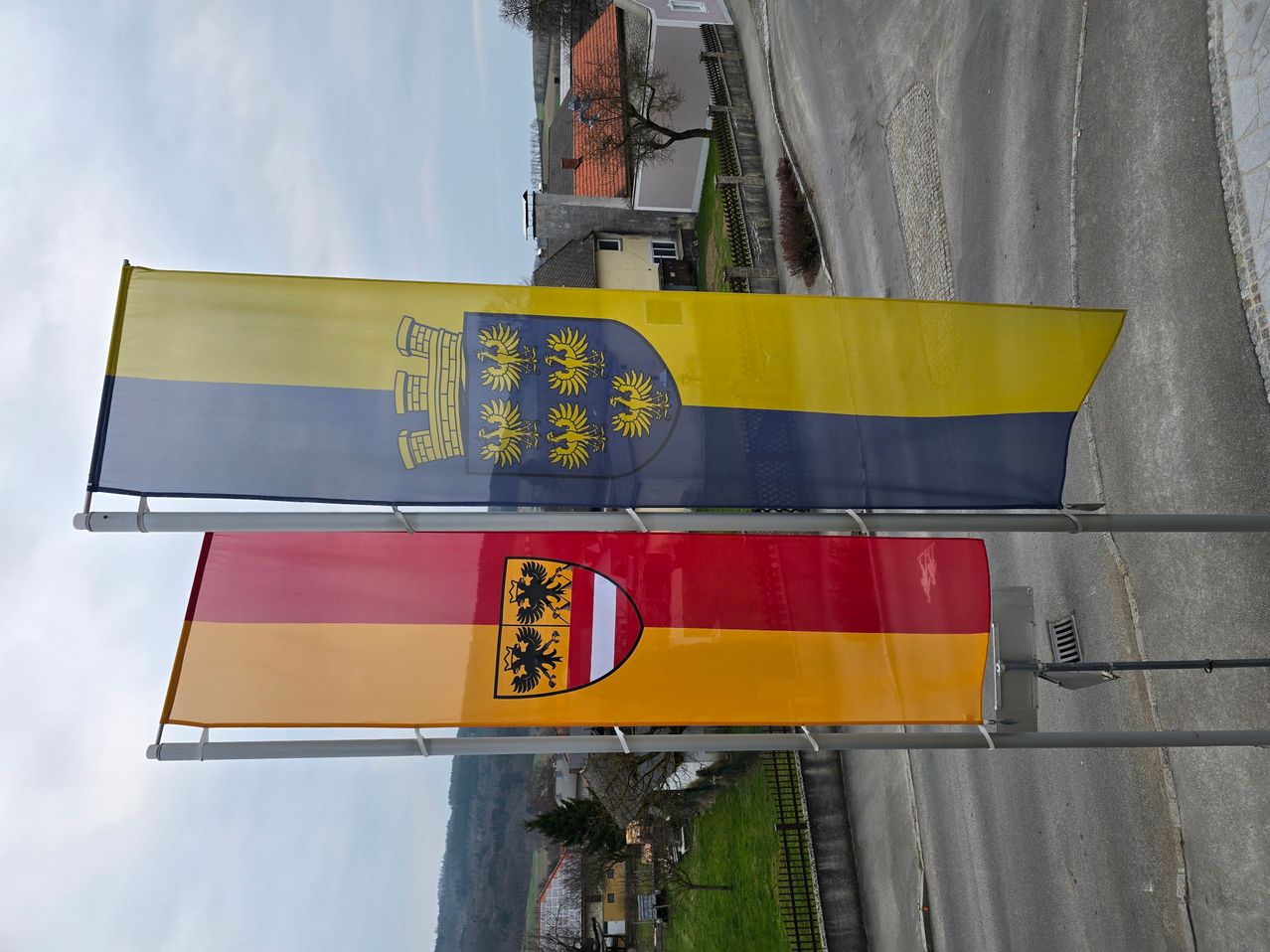 Zwei Banner sind im Freien ausgestellt. Eines ist ein blau-gelbes Banner mit einem goldenen Löwen- und Schlosswappen. Das andere ist ein rot-gelbes Banner mit einem schwarzen Adlerwappen in der Mitte.