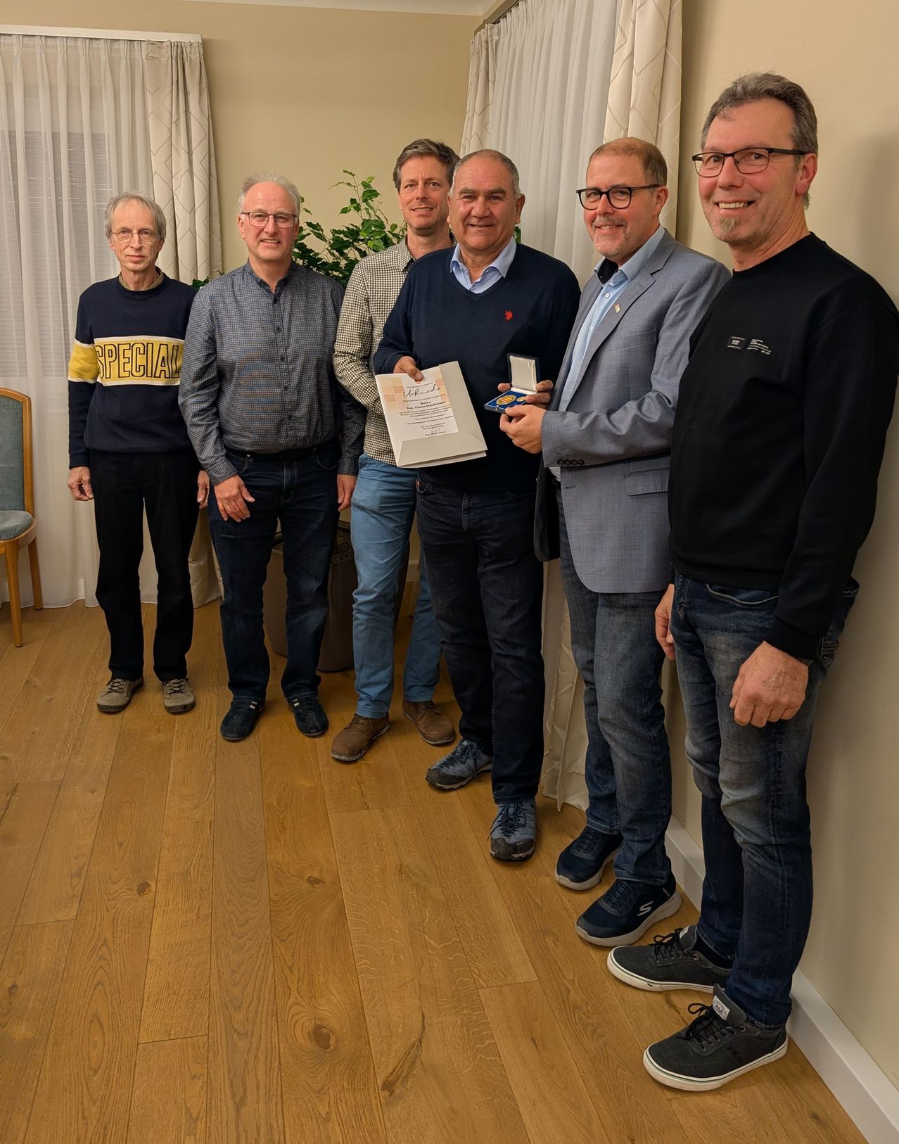 Fünf Männer stehen auf einem Holzboden. Sie tragen Freizeitkleidung und scheinen für ein Foto zu posieren. Ein Mann hält ein Zertifikat.