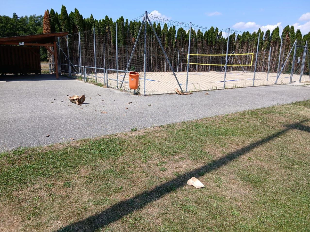 Ein leerer Außensportplatz mit einem Volleyballnetz und einem Mülleimer. Der Platz ist von einem Metallzaun umgeben.