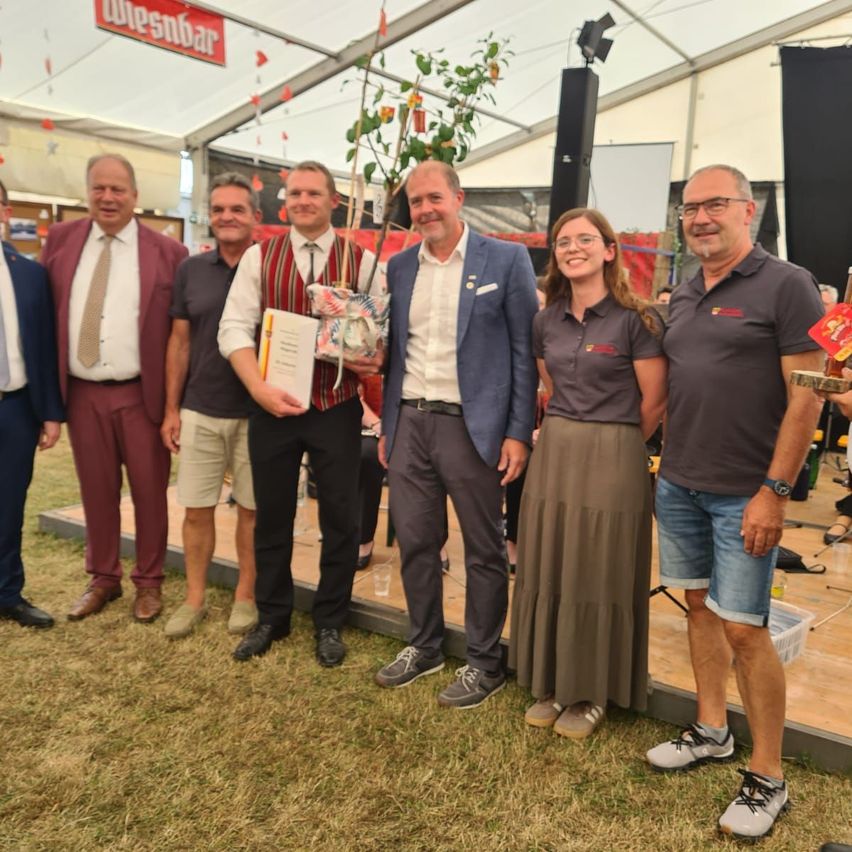 Eine Gruppe von Menschen, darunter Männer und Frauen, steht zusammen auf einer Bühne mit einem Hintergrund aus einem Zelt und einem Schild mit der Aufschrift 'Wiesenbar'. Sie lächeln und scheinen für ein Foto zu posieren.