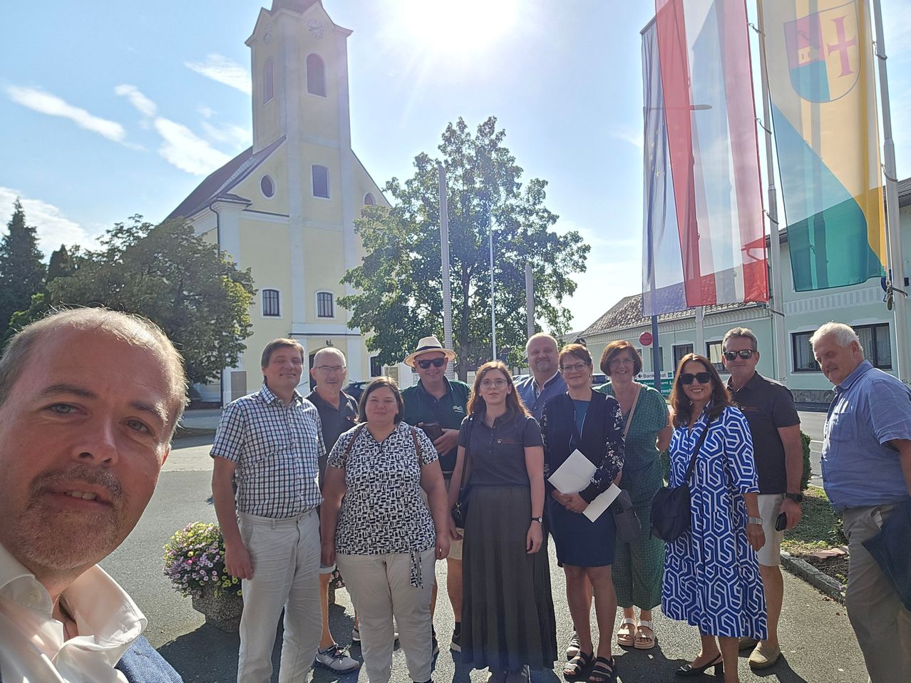 Eine Gruppe von Menschen steht vor einer Kirche mit Turm und Fahnen an einem sonnigen Tag.