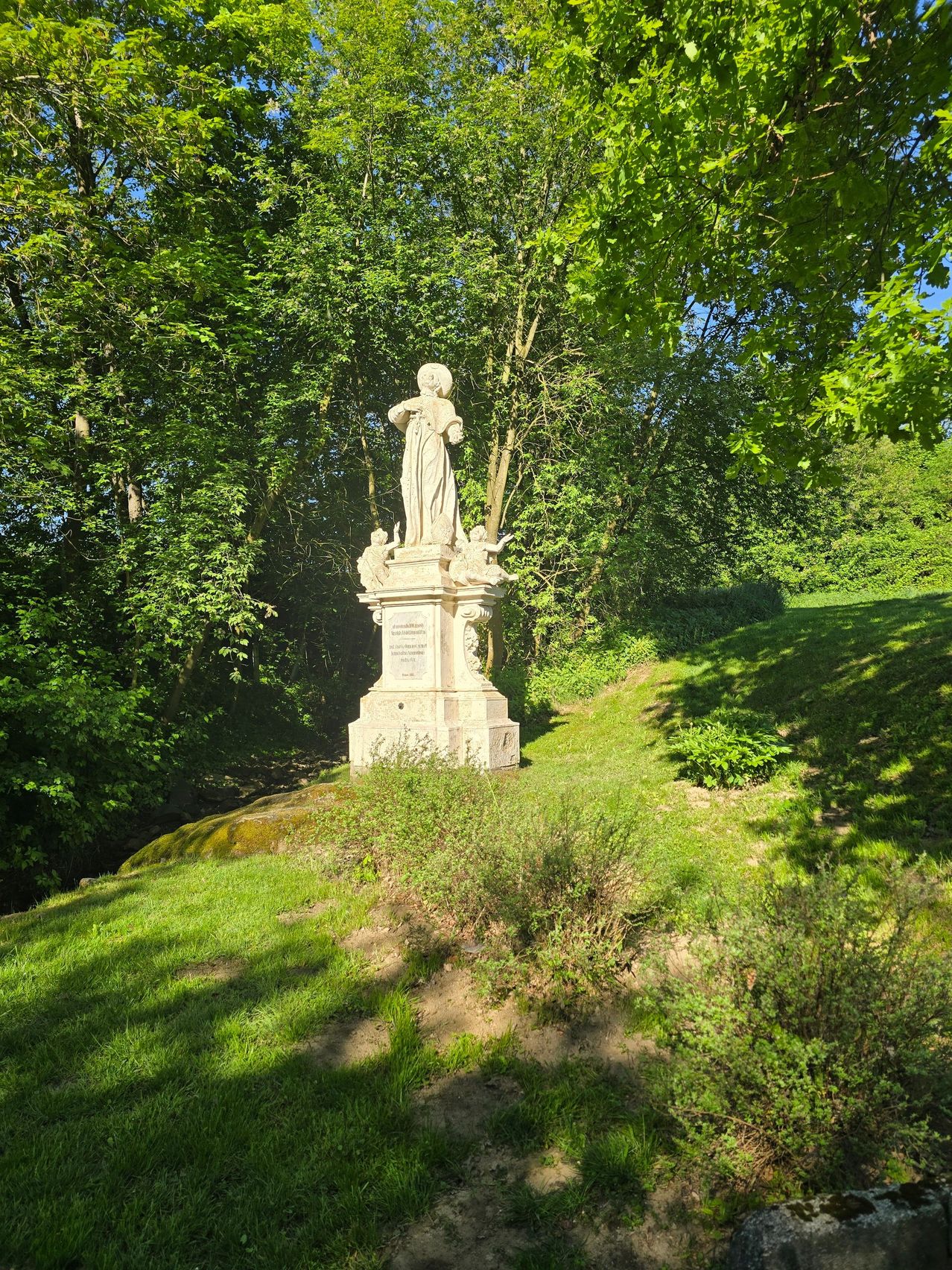 Eine Marmorstatue einer Frau steht auf einem Hügel, umgeben von üppigem Grün und Bäumen. Die Statue wird vom Sonnenlicht beleuchtet, das Schatten auf das Grasland wirft.