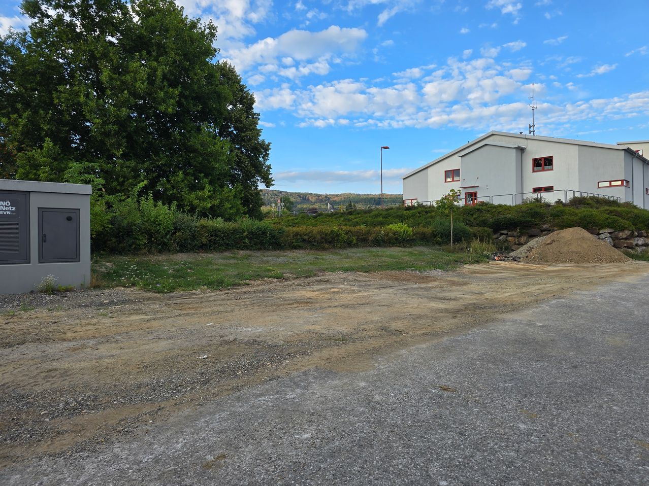 Ein leeres Grundstück mit Kies und Gras, ein weißes Gebäude mit roten Fenstern im Hintergrund und eine Straßenlaterne.