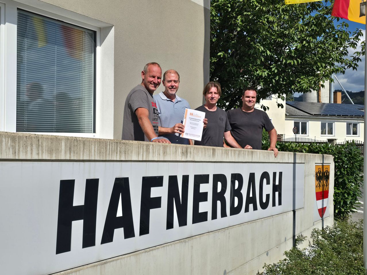 Vier Männer stehen vor einem Schild, auf dem 'Hafnerbach' steht. Der Mann in der Mitte hält ein Zertifikat. Dahinter ist ein Fenster, ein Baum und ein Haus.