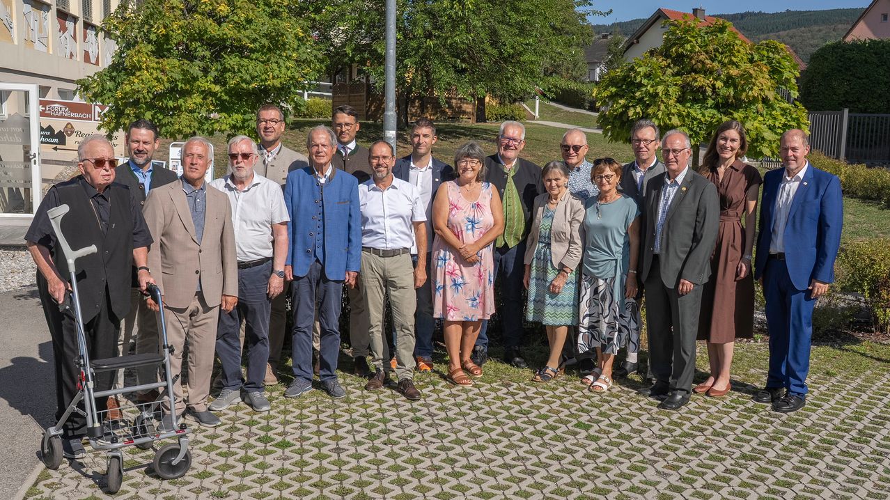 Eine Gruppe von Menschen, die formell gekleidet sind, steht zusammen in einem Park. Sie lächeln und scheinen für ein Foto zu posieren. Im Hintergrund befinden sich Bäume und ein Haus.