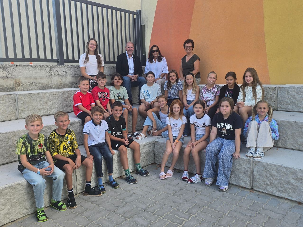 Eine Gruppe von Kindern und Erwachsenen posiert für ein Foto auf einer Steinbank vor einer farbenfrohen Wand.