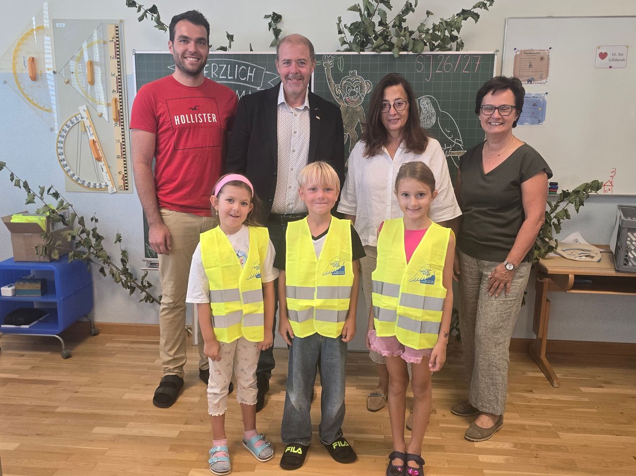 Eine Gruppe von Erwachsenen und Kindern, darunter zwei Jungen in gelben Westen, stehen gemeinsam vor einer Tafel. Die Erwachsenen lächeln und scheinen für ein Foto zu posieren.