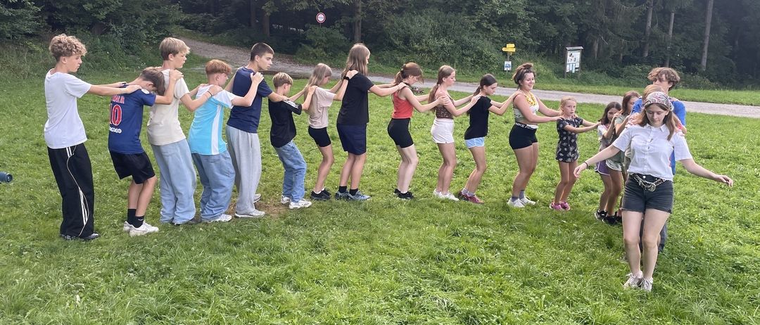 Eine Gruppe von Kindern und Jugendlichen bildet einen Kreis auf einer Wiese, wobei sie sich an den Händen halten. Sie sind lässig gekleidet. Im Hintergrund befinden sich Bäume und eine Straße.