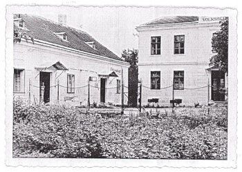 Ein Schwarz-Weiß-Foto zeigt zwei Gebäude, eines mit einem Garten und einem Zaun und das andere mit einem Schild, das 'Volkschule' liest.