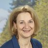 Eine Frau mit blondem Haar lächelt, möglicherweise für ein Foto. Sie trägt einen blauen Blazer über einem gemusterten Oberteil. Der Hintergrund ist unscharf, mit Bäumen und blauem Himmel.