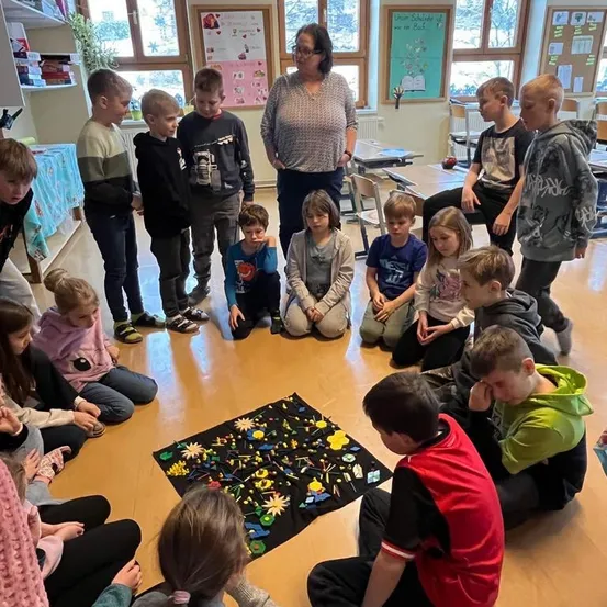 Eine Gruppe von Kindern und eine Frau sind in einem Klassenzimmer, einige sitzen auf dem Boden um eine Matte mit verschiedenen Gegenständen herum. Die Frau steht in der Mitte.