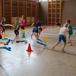 Kinder spielen mit Hula-Hoops und Kegeln in einer Turnhalle. Sie laufen und navigieren durch die Hindernisse. Einige beugen sich, während andere stehen.