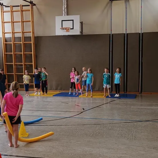 Mehrere Kinder sind in einer Turnhalle versammelt. Sie stehen auf blauen Matten. Einige Kinder tragen Turnschuhe. An der Wand ist ein Basketballkorb zu sehen.