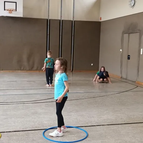 In einer Turnhalle steht ein Mädchen mit einem Hula-Hoop, während zwei Kinder auf einem Skateboard spielen. Die Wände sind mit Basketball-Ausrüstung geschmückt.