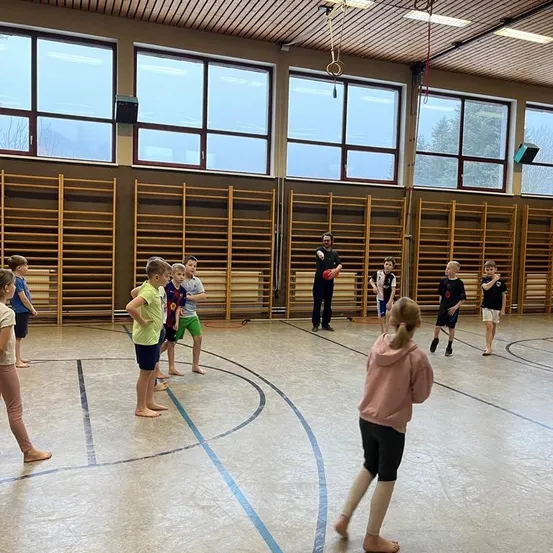 In einer Turnhalle nehmen mehrere Kinder an einer Aktivität teil, möglicherweise einem Spiel oder einer Übung, die von einem Mann beaufsichtigt wird. Holzregale säumen die Wände.
