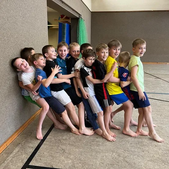 Eine Gruppe junger Jungen steht eng beieinander und lächelt in einer Turnhalle. Sie sind barfuß und tragen verschiedenfarbige T-Shirts.
