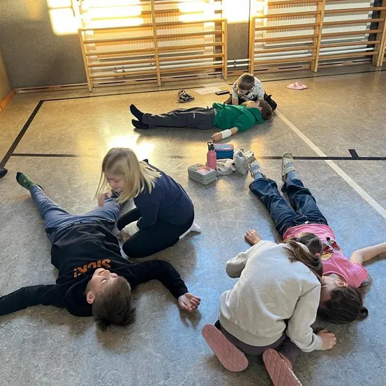 Mehrere Kinder liegen auf dem Boden in einem Raum, wobei ein Mädchen einen Jungen untersucht. Ein anderes Kind liegt auf einer Matte mit einem Kissen und einem Verband.