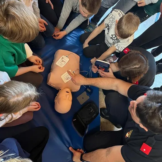 Kinder beobachten, wie ein Mann in Uniform CPR an einem Mannequin demonstriert, mit einem AED in der Nähe.