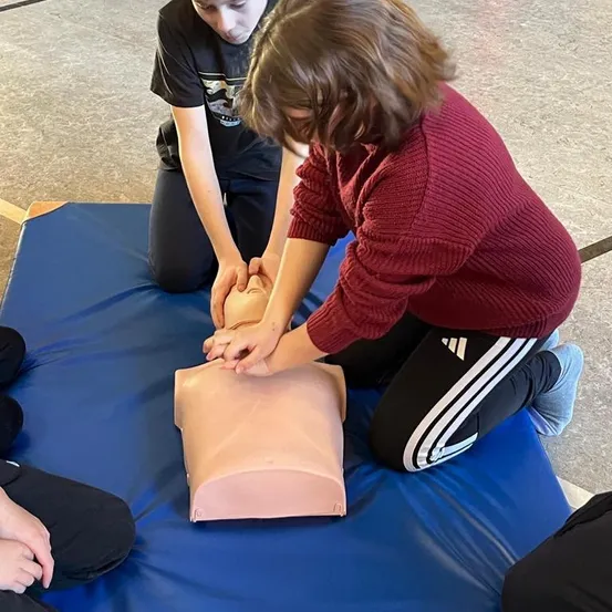 Zwei Kinder üben in einer Turnhalle CPR an einem Dummies, wobei ein Kind auf die Brust des Dummies drückt.