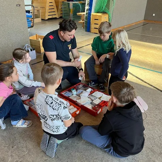 Eine Gruppe von Kindern, darunter Jungen und Mädchen, sitzen auf dem Boden und lauschen einem Mann in Uniform. Sie scheinen eine Sicherheitsschulung zu absolvieren.