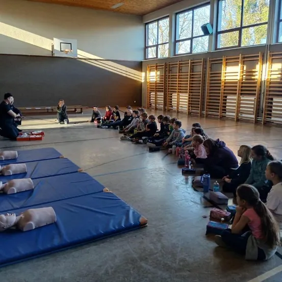 Eine Gruppe von Schülern sitzt in einem Kreis um einen Ausbilder mit Trainingsmanequins auf dem Boden in einer Turnhalle.