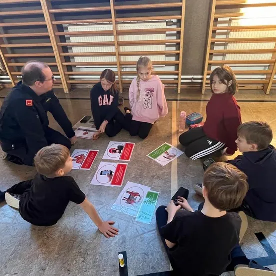 Ein Mann und mehrere Kinder sitzen um einen Tisch mit Informationskarten auf dem Boden. Der Mann schaut die Kinder an. Die Kinder beschäftigen sich mit den Karten und dem Mann.