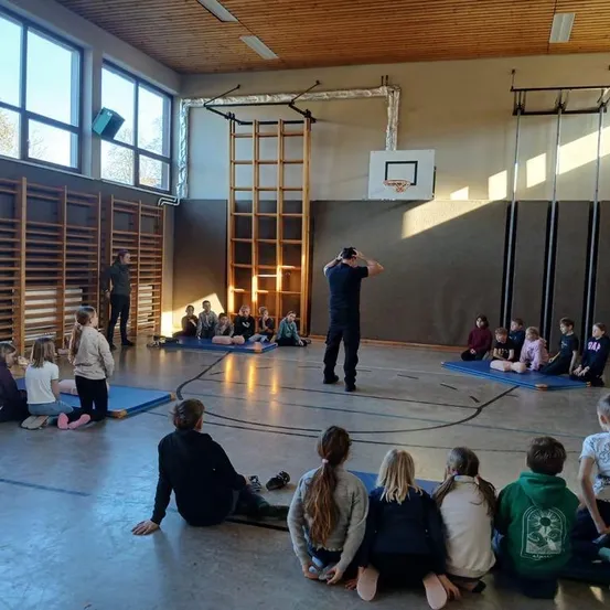 In einer Turnhalle sitzen Kinder in einem Kreis auf blauen Matten. Eine Person steht in der Mitte, und eine andere steht an der Seite. Der Raum hat Holzregale, Fenster und einen Basketballkorb.
