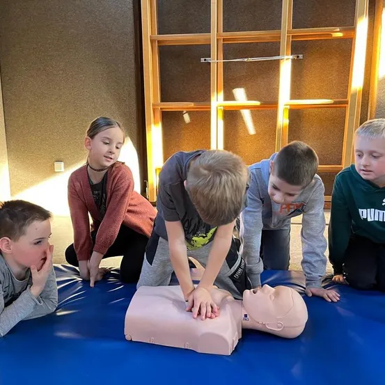 Fünf Kinder üben CPR an einem Mannequin. Ein Kind drückt auf die Brust des Dummies, während ein anderes zusieht. Dahinter befindet sich ein hölzernes Regal mit einem Kreuz.