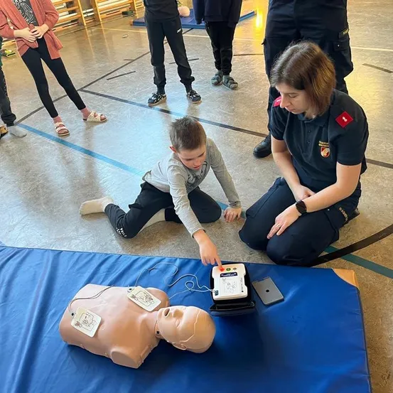 Eine Frau und ein Junge üben CPR an einer Puppe. Sie sind von anderen umgeben, die in einer Turnhalle beobachten.