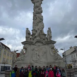 Eine Gruppe von Kindern und Erwachsenen steht an einem bewölkten Tag vor einer großen Statue. Die Statue ist detailliert mit Skulpturen von Figuren und hat Lampen angebracht.