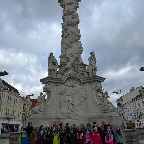 Eine Gruppe von Kindern und Erwachsenen steht an einem bewölkten Tag vor einer großen Statue. Die Statue ist detailliert mit Skulpturen von Figuren und hat Lampen angebracht.