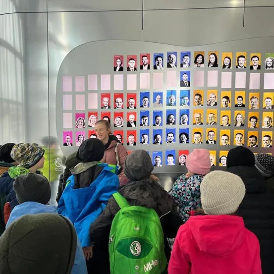 Eine Gruppe von Menschen, wahrscheinlich Schüler, versammelt sich vor einer großen Wand, die mit Fotos verschiedener Personen bedeckt ist. Eine Frau in der Mitte trägt eine Brille und eine Handtasche.