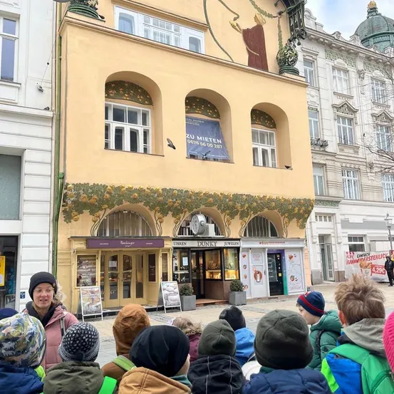 Eine Gruppe von Kindern und einer erwachsenen Frau steht vor einem gelben Gebäude mit einem Wandbild und einem Banner, das 'Zu Mieten' besagt.