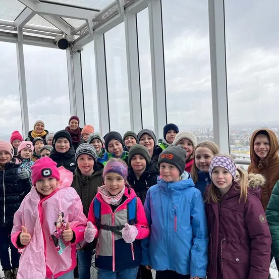 Eine Gruppe von Kindern in Winterkleidung posiert für ein Foto in einem Aussichtsbereich mit Glasfenstern. Die Kinder lächeln alle.