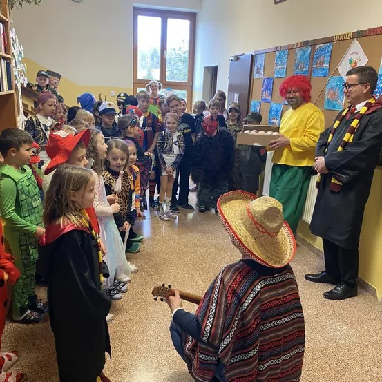 Eine Gruppe von Kindern in Kostümen ist in einem Raum versammelt, wobei ein Mann eine Schachtel mit Eiern hält. Ein anderer Mann im Sombrero spielt Gitarre.