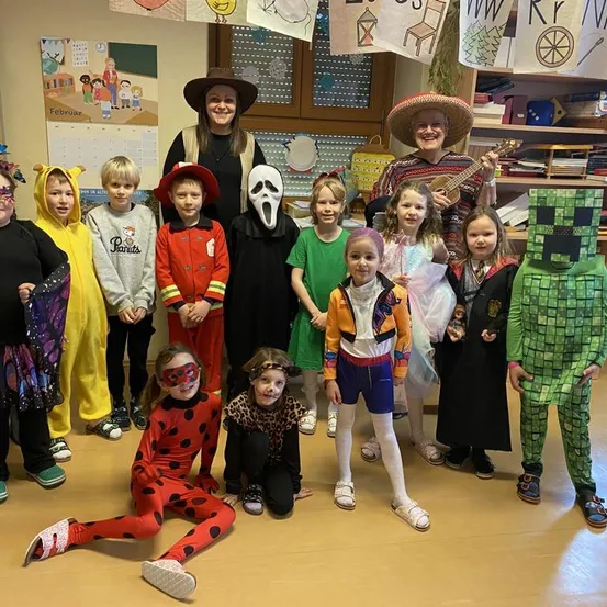 Eine Gruppe von Kindern und Erwachsenen in Kostümen posiert für ein Bild in einem Klassenzimmer. Verschiedene Kostüme umfassen eine Marienkäferin, einen Vogelscheuche und einen Minecraft-Charakter.