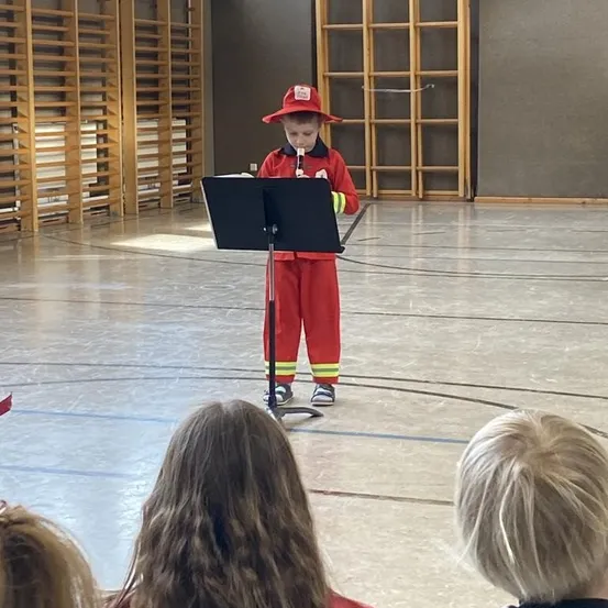 Ein kleiner Junge, verkleidet als Feuerwehrmann, steht an einem Rednerpult in einer Turnhalle und hält ein Mikrofon in der Hand. Mehrere Kinder beobachten ihn aufmerksam.
