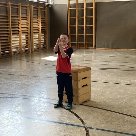 Ein Junge lächelt und posiert in einer Turnhalle, neben einer Holzkiste stehend. Hinter ihm befinden sich Holzregalen und eine Wand mit einem Fenster.