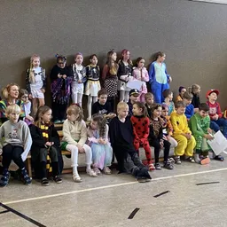 Eine Gruppe von Kindern in verschiedenen Kostümen sitzt auf Bänken in einer Halle. Einige halten Papiere, und einige stehen hinter ihnen.