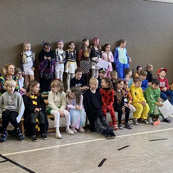 Eine Gruppe von Kindern in verschiedenen Kostümen sitzt auf Bänken in einer Halle. Einige halten Papiere, und einige stehen hinter ihnen.