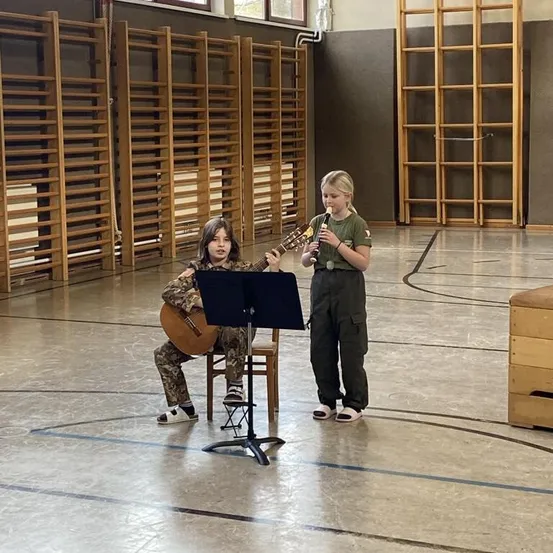 Zwei Kinder spielen ein Musikstück in einer Turnhalle. Eines spielt eine Flöte, während das andere eine Gitarre spielt. Sie stehen vor einem Notenständer.