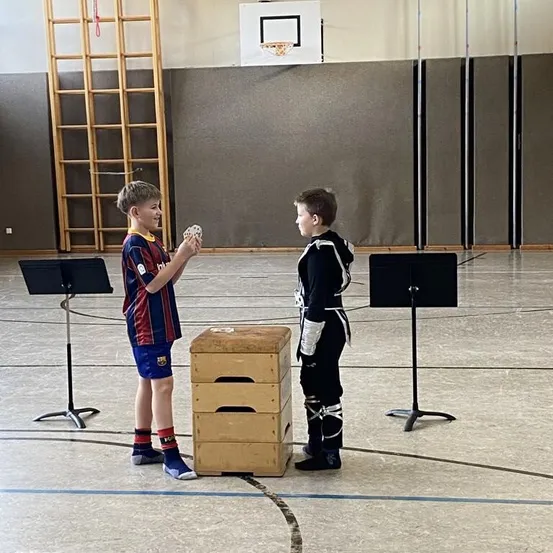 Zwei Jungen stehen in einer Turnhalle. Einer hält Karten, der andere sieht sie an. Musikständer stehen hinter ihnen. Eine hölzerne Leiter ist links. Ein Basketballkorb ist an der Wand montiert.