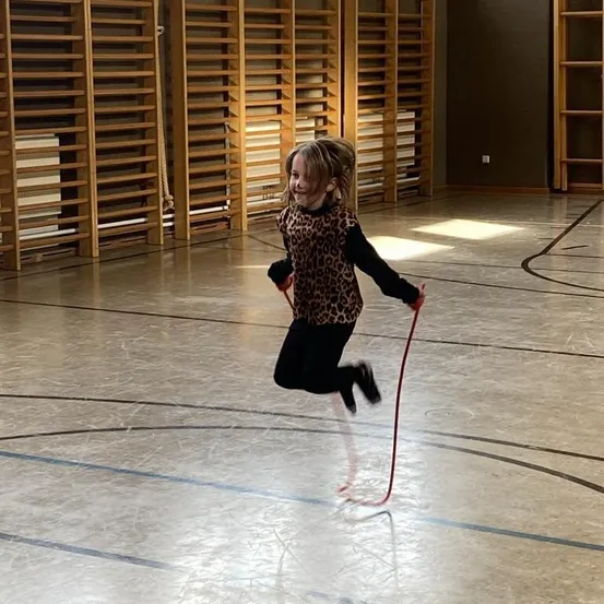 Ein kleines Mädchen springt Seil in einer Turnhalle mit Holzregalen im Hintergrund. Sie trägt ein Leopardenmuster-Top und schwarze Leggings.