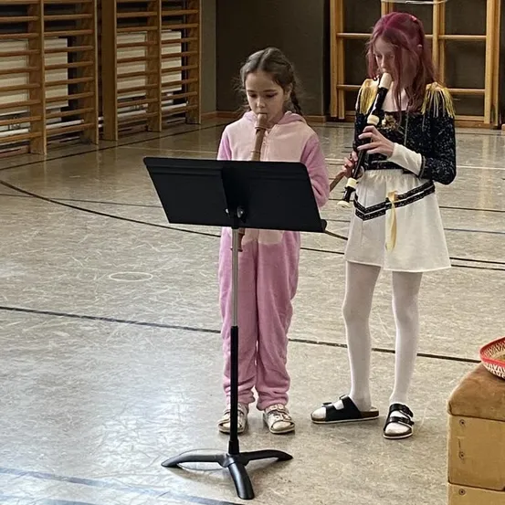 Zwei Mädchen spielen Flöten auf der Bühne, eine in rosa Schlafanzug, die andere in weißem Kleid mit roter Perücke. Ein Notenständer und ein Korb sind in der Nähe.