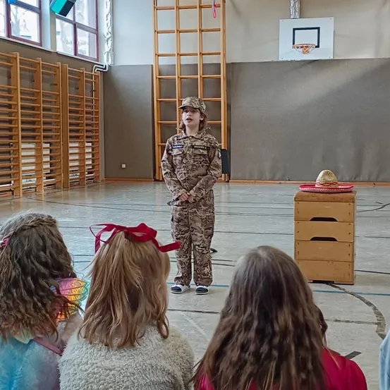 Ein junger Mensch in einer Tarnuniform steht in einem Raum mit mehreren Kindern, die zuschauen. Der Raum hat ein Basketballnetz und Holzlager.