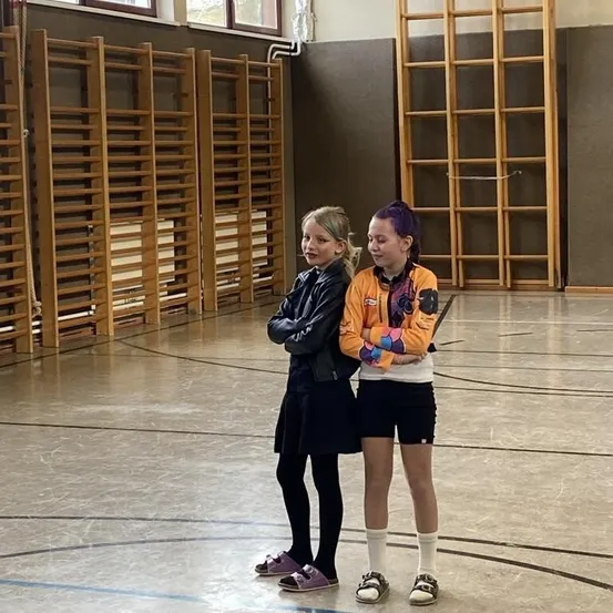 Zwei Mädchen stehen in einer Turnhalle, eine mit lila Haaren und das andere mit blond. Sie tragen Turnschuhe. Dahinter befinden sich Holzleitern und Ausrüstung.