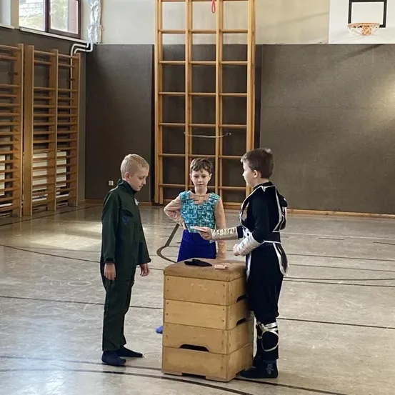 Drei Kinder in einer Turnhalle, eines gibt dem anderen eine Karte, stehend bei einem Holzkisten. Ein Kind trägt ein Kostüm mit Rüstung.