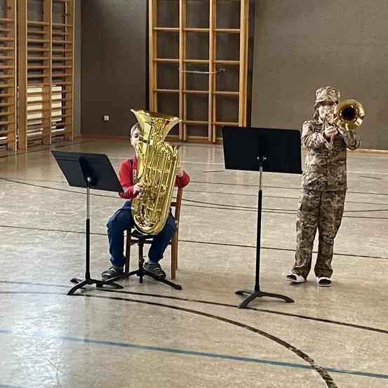 Zwei Kinder spielen Blechblasinstrumente in einem Raum. Eins sitzt auf einem Stuhl mit einer Tuba, das andere steht mit einer Posaune. Sie tragen beide Tarnkleidung.